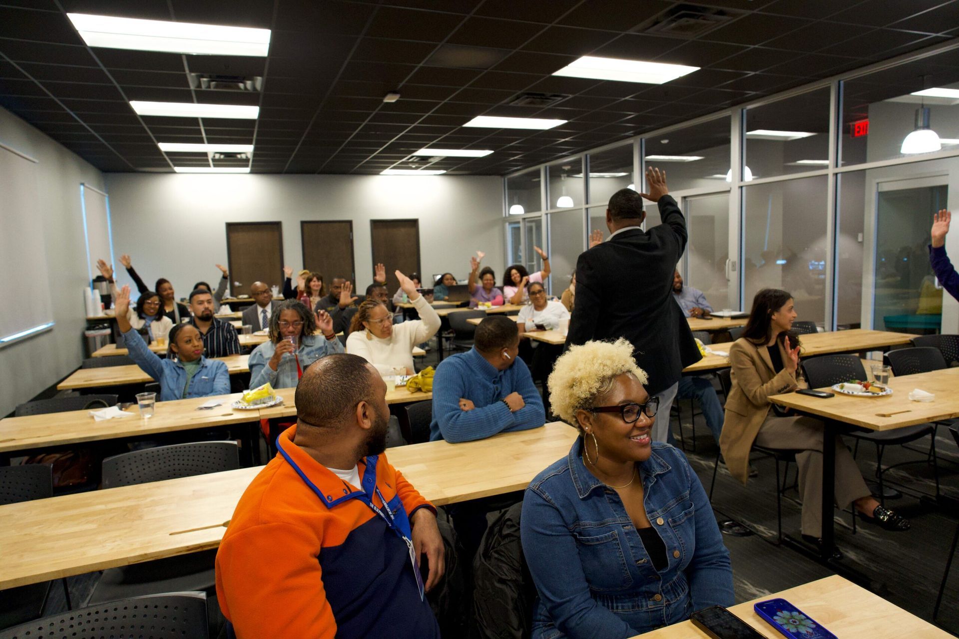 Meeting room full of seated people listening to a speaker at the front, with tables and glass walls.