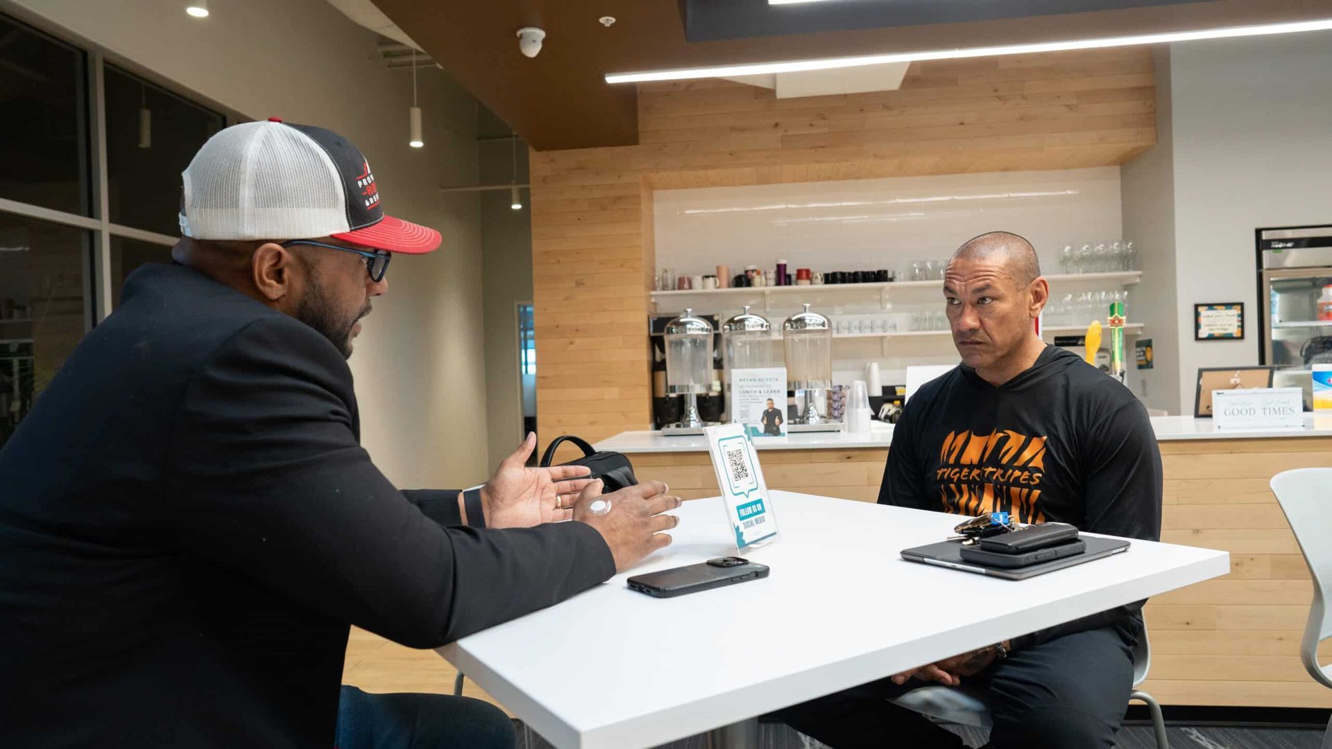 Two men talking across a table in a bright office break room, with phones and a drink cup on the table