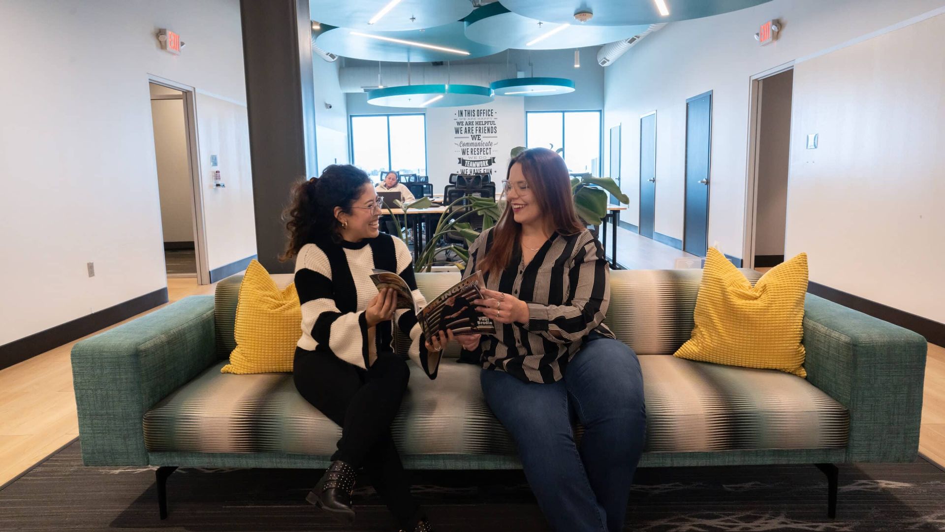 Two people talking on a green couch in a bright hallway lounge with yellow pillows.