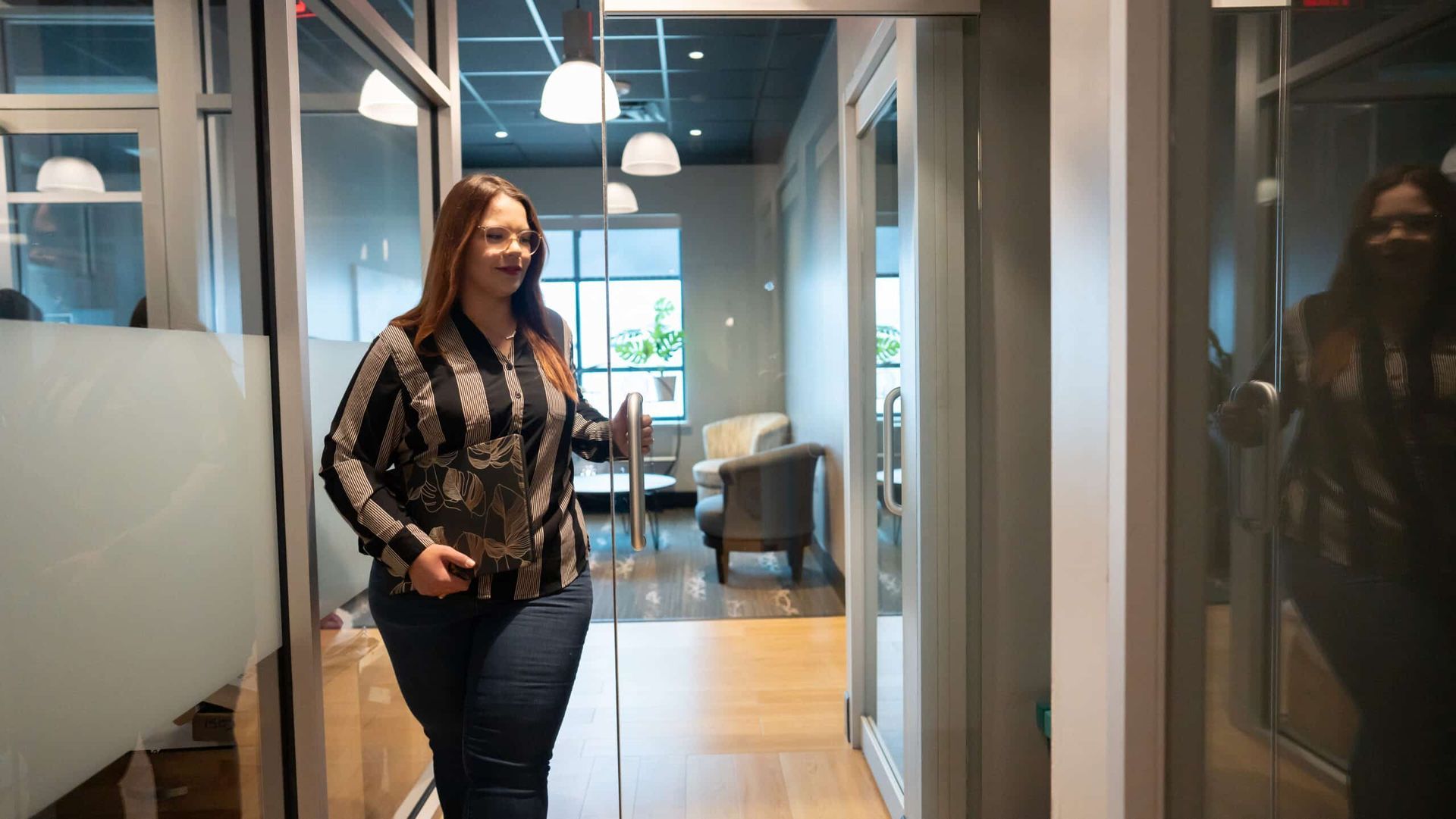 Woman walking through a modern office hallway, wearing a patterned sweater and jeans.