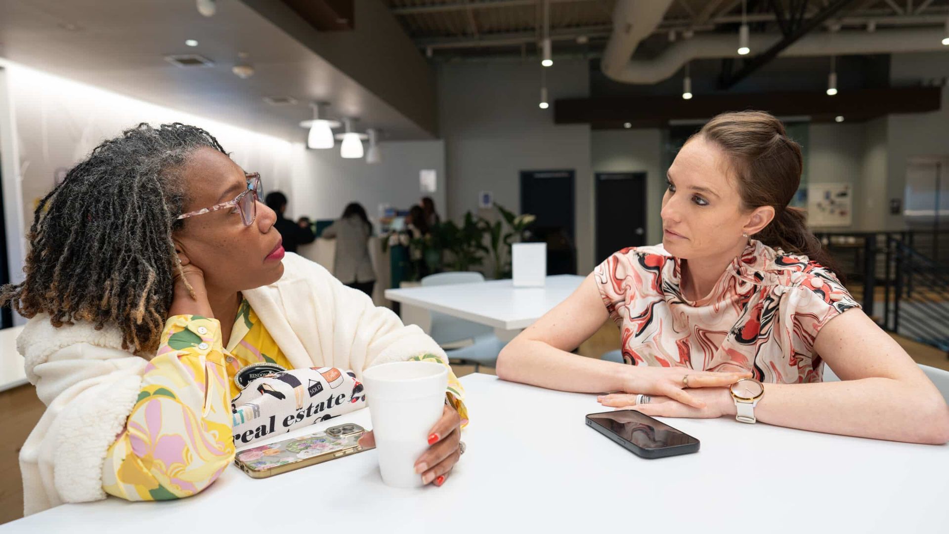 Two people talking across a table in a modern office, with papers and a phone between them.