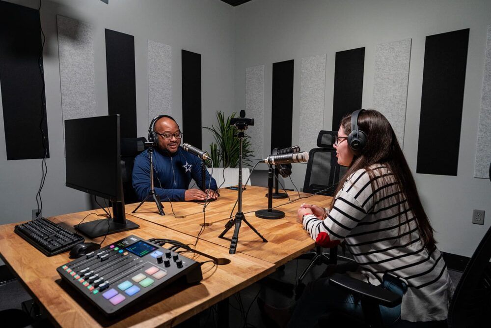 Podcast studio with two people speaking across microphones at a wooden table, with audio mixer and monitors.