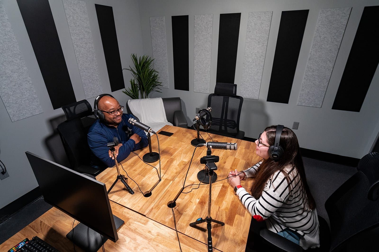 Two people speaking into microphones in a podcast recording studio with a wooden table and acoustic panels.