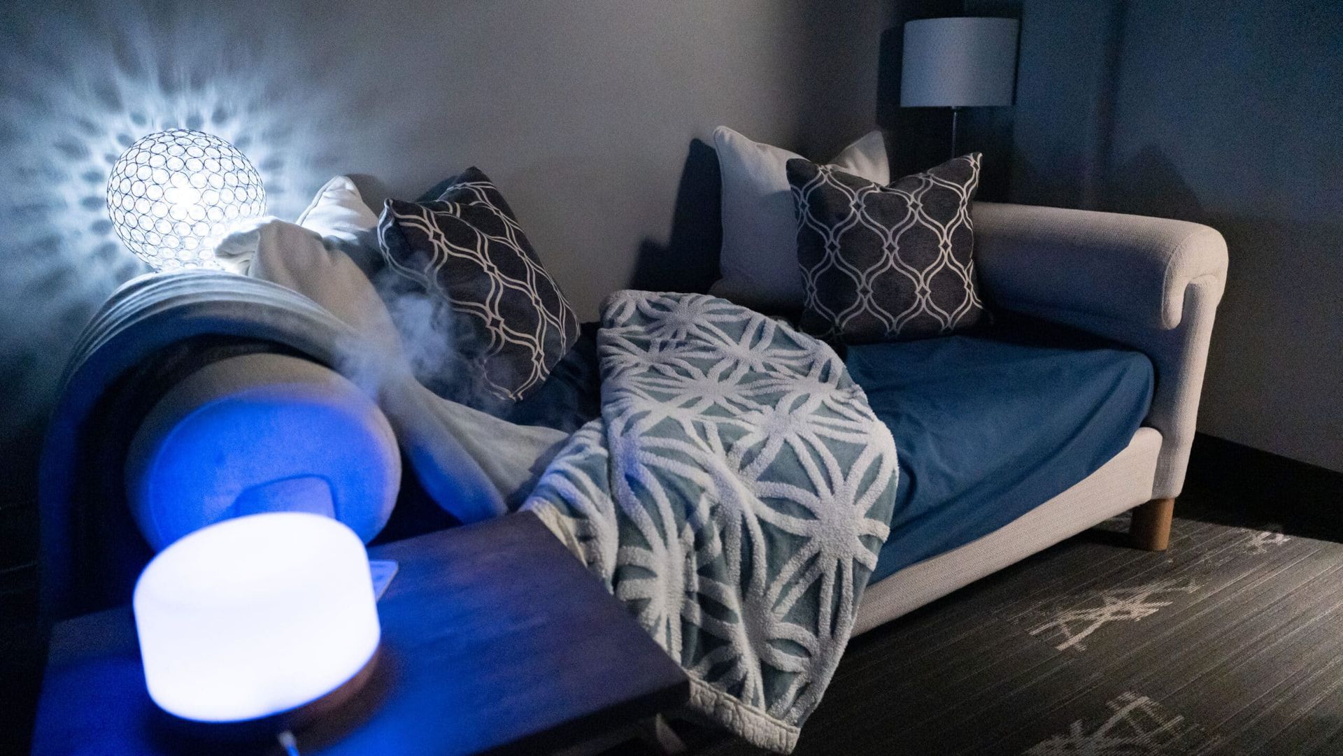 Cozy living room with a blue sofa, patterned pillows, a throw blanket, and soft glowing lamps