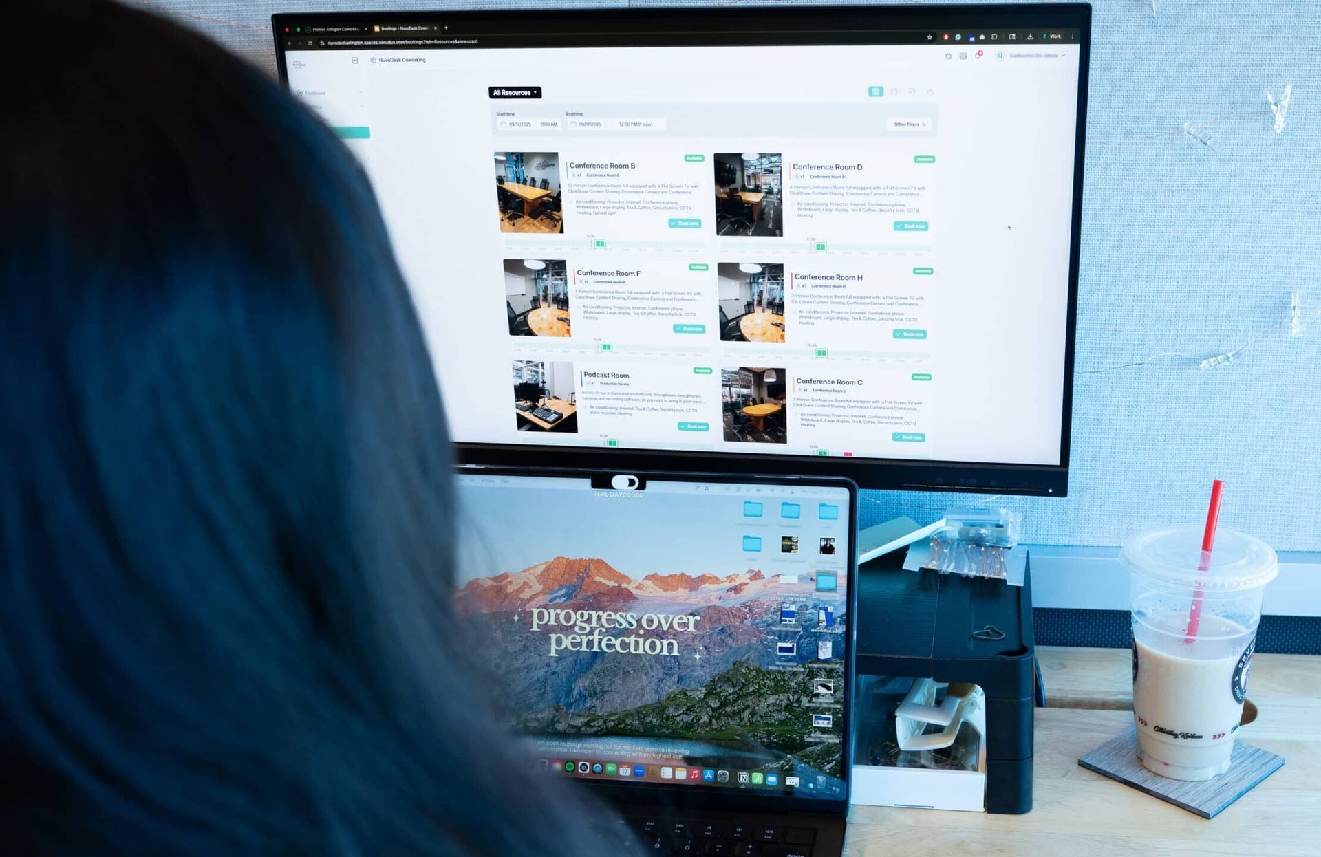 Person using a laptop beside a desktop monitor showing a website, with a drink on the desk.