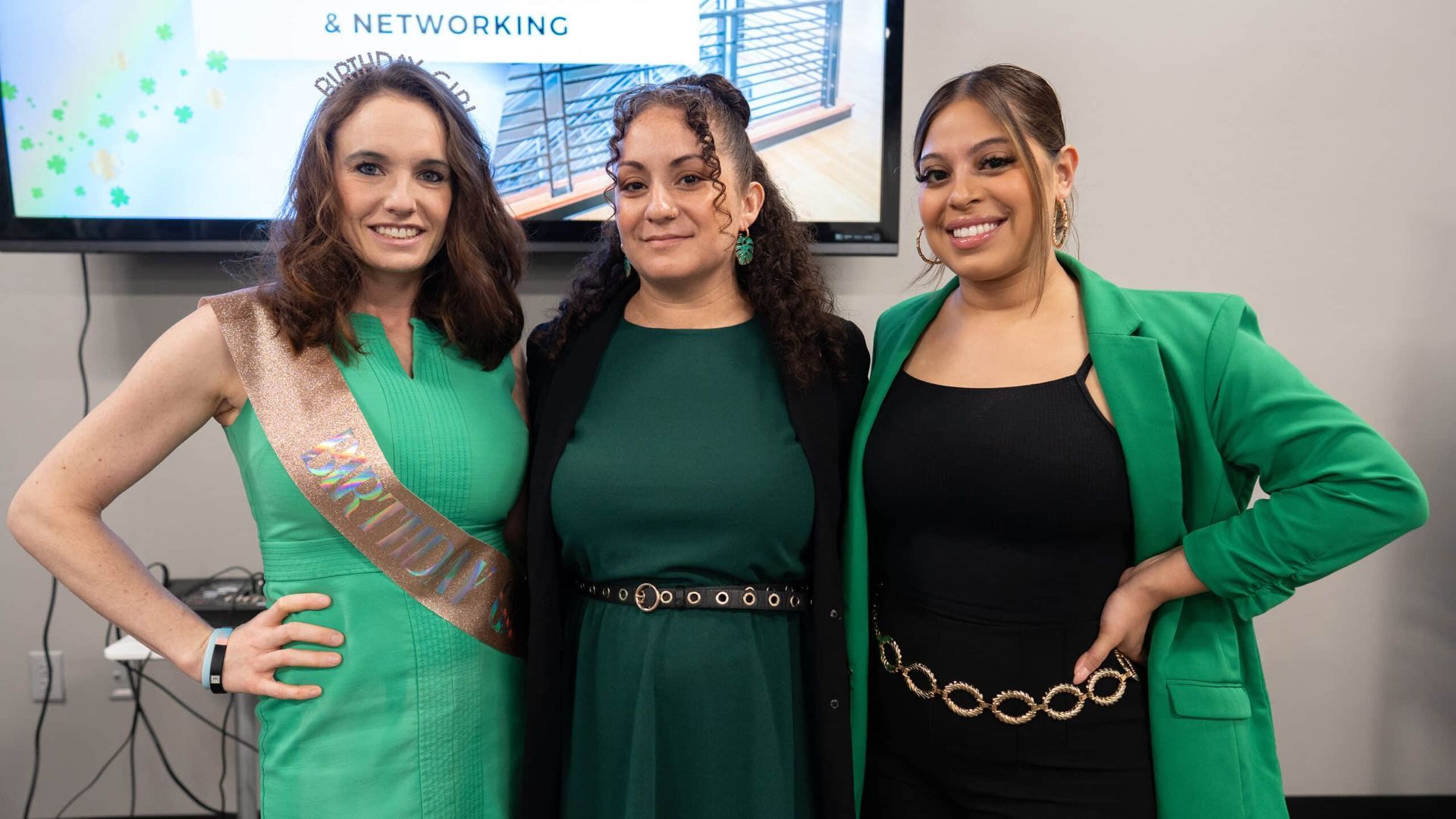 Three people in green and black outfits posing at a networking event, smiling in front of a screen.