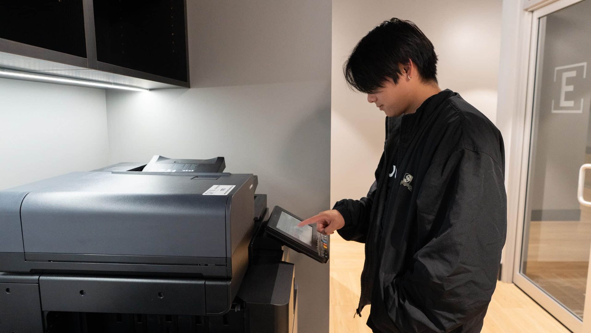 Man using a printer/copier in an office hallway, holding the control panel while the machine prints.