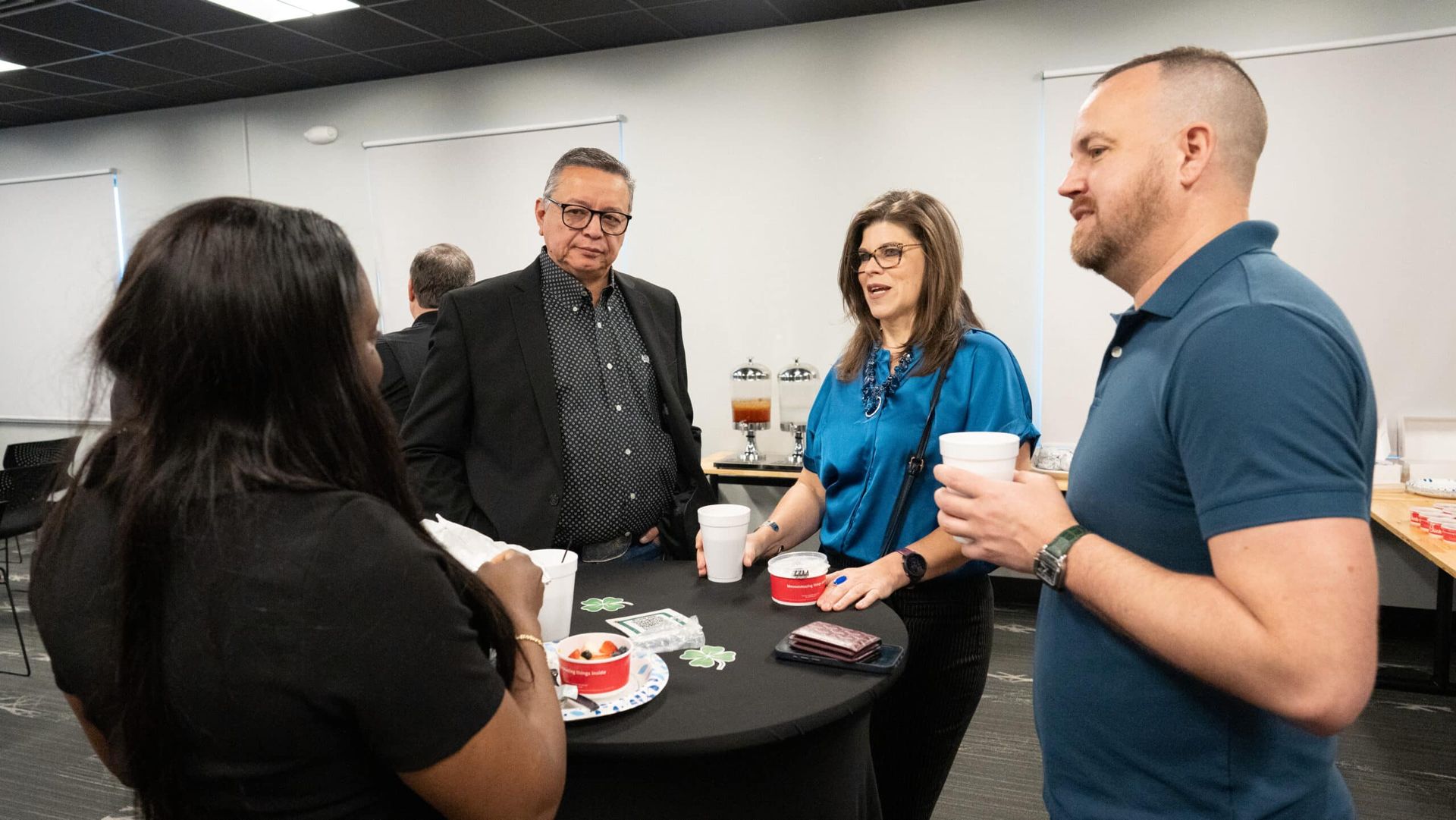 Four people stand chatting around a high table with snacks in a bright office break room.