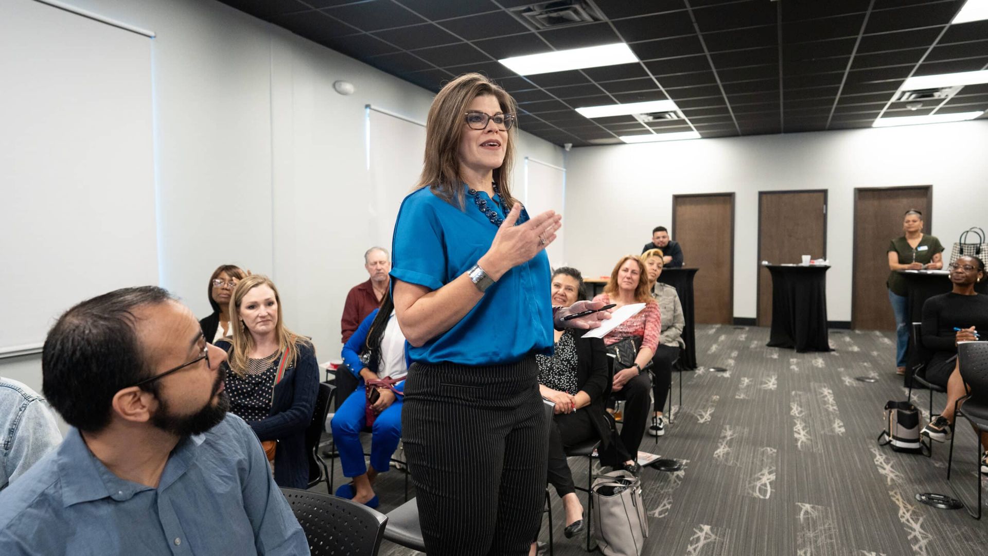 Woman speaking to a seated audience in a conference room, holding a microphone and smiling.