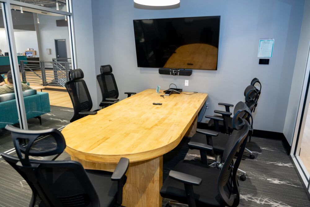 Modern conference room with a long wooden table, black office chairs, and a wall-mounted TV.