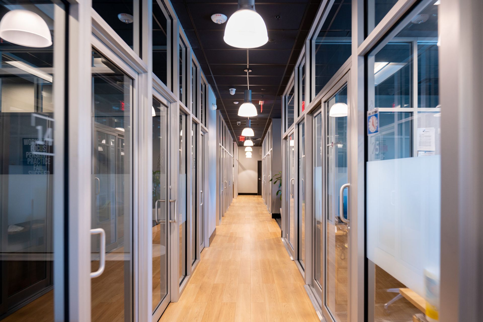 Modern office hallway with glass-walled rooms, wood floor, and hanging lights
