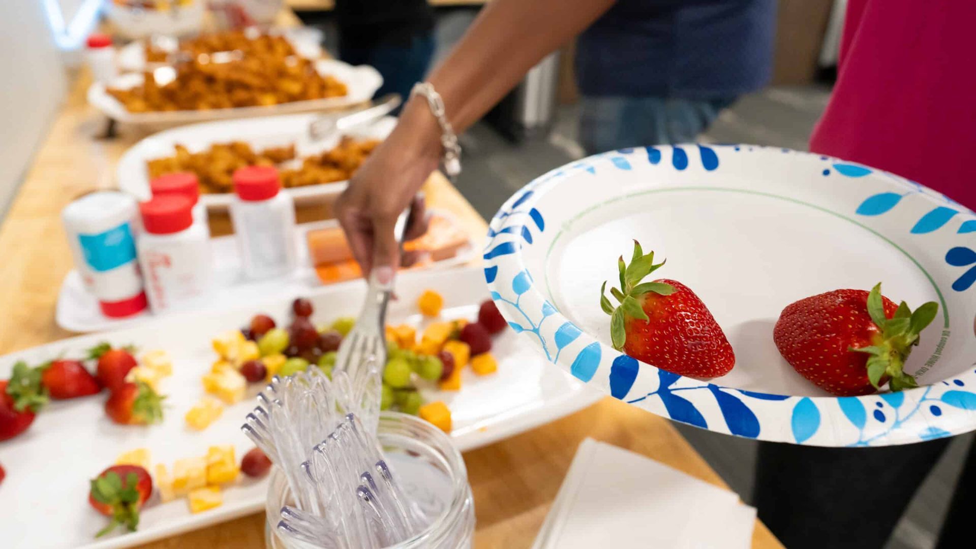 Hands serving strawberries onto a paper plate at a dessert buffet table