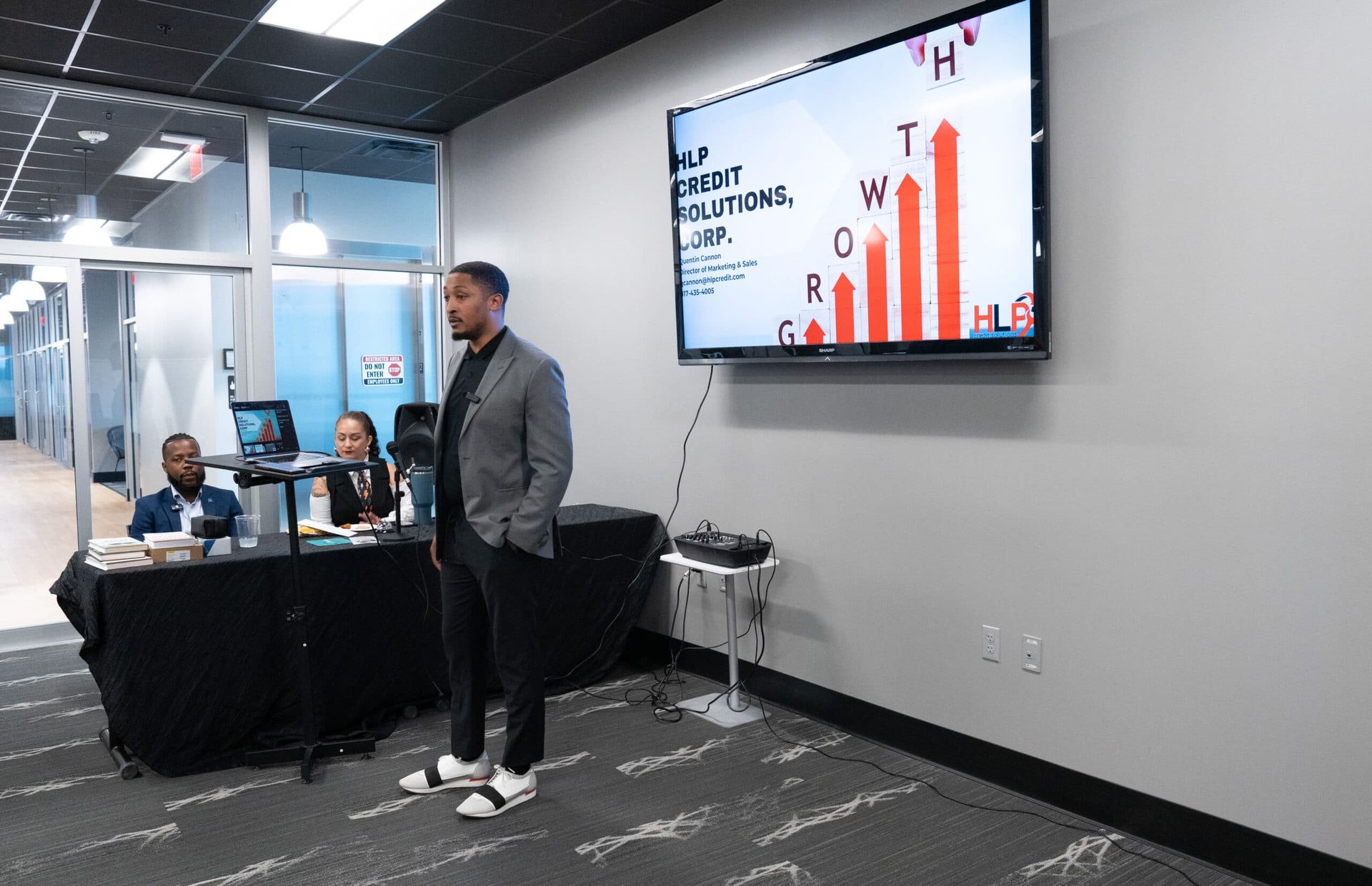 Presenter speaking beside a wall-mounted screen in a modern office meeting room with seated attendees