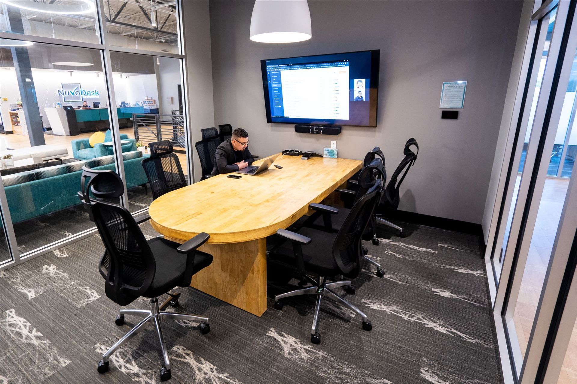 Conference room with oval wood table, black chairs, wall-mounted screen, and office desk setup