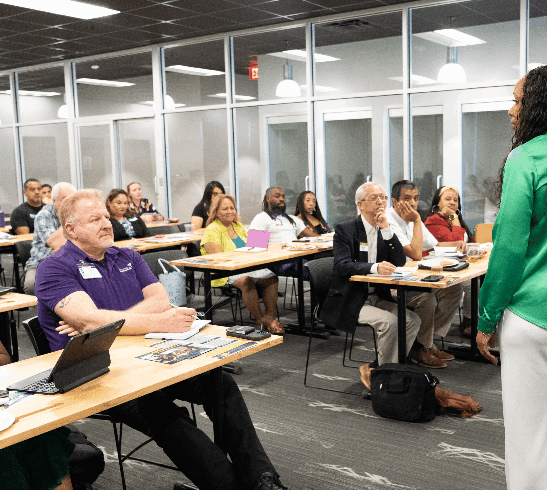 People attending a classroom presentation, seated at desks and listening to a speaker in a green jacket