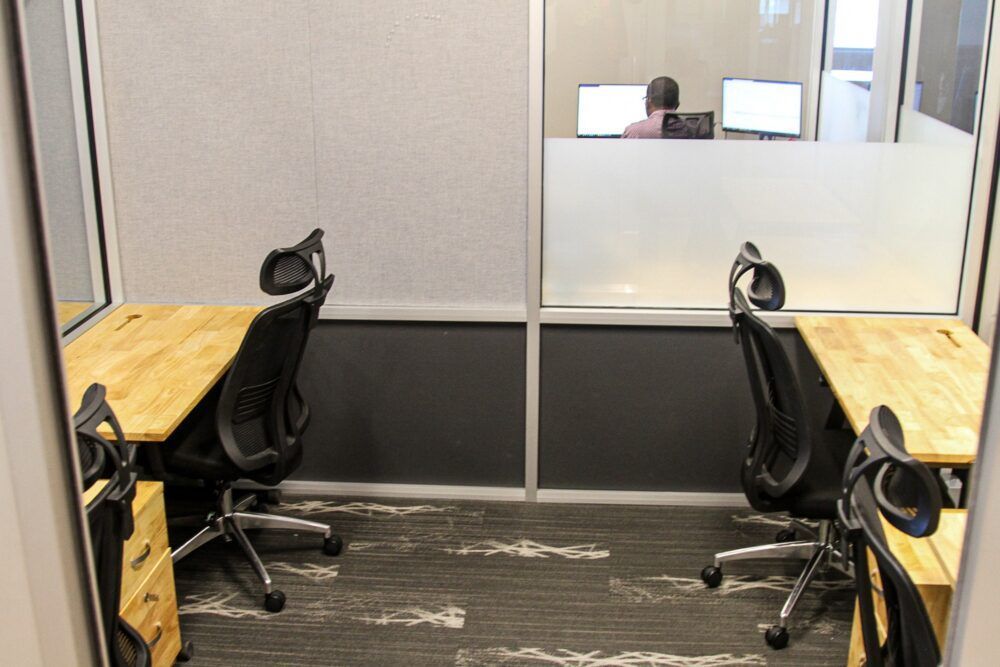 Small office with two wooden desks, black chairs, and a frosted glass partition.