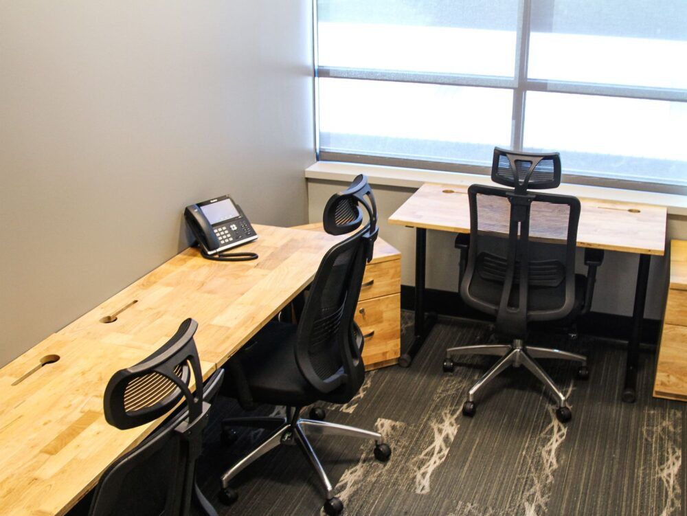 Small office with two desks, rolling chairs, and desk phones beside a window.