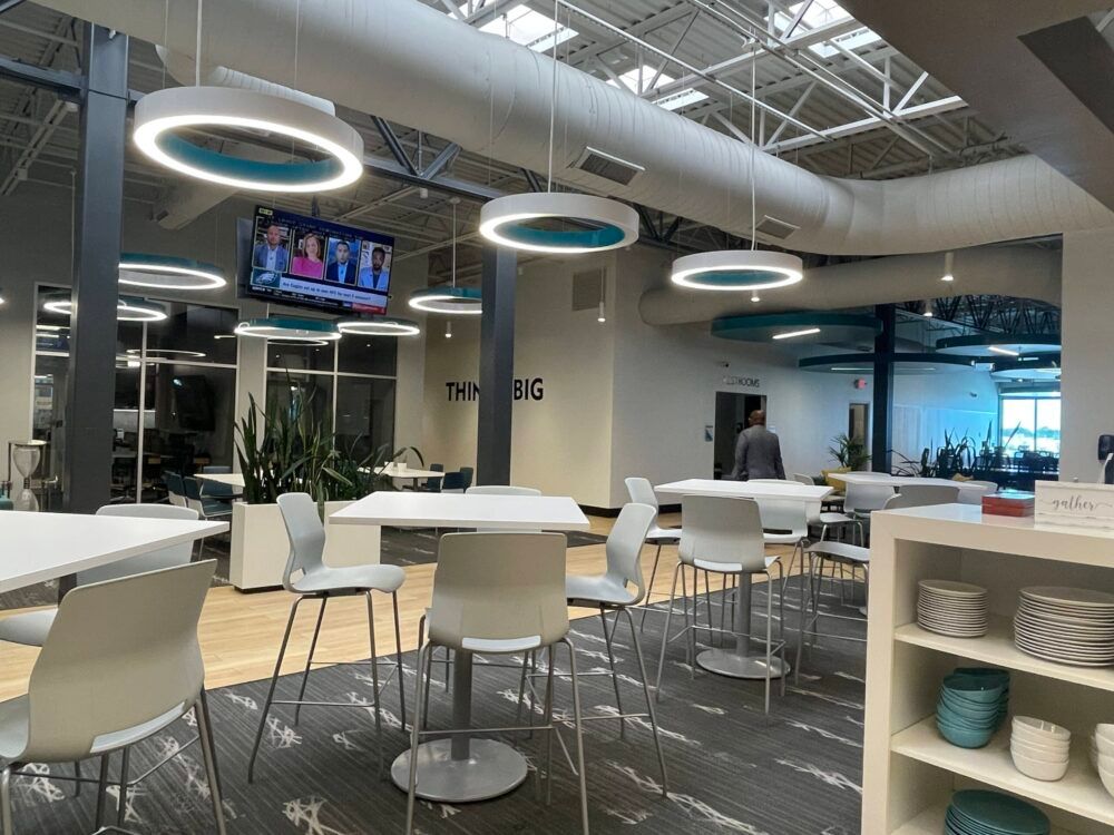 Modern lounge with white tables and chairs, hanging ring lights, TV screens, and shelves in a bright open office.