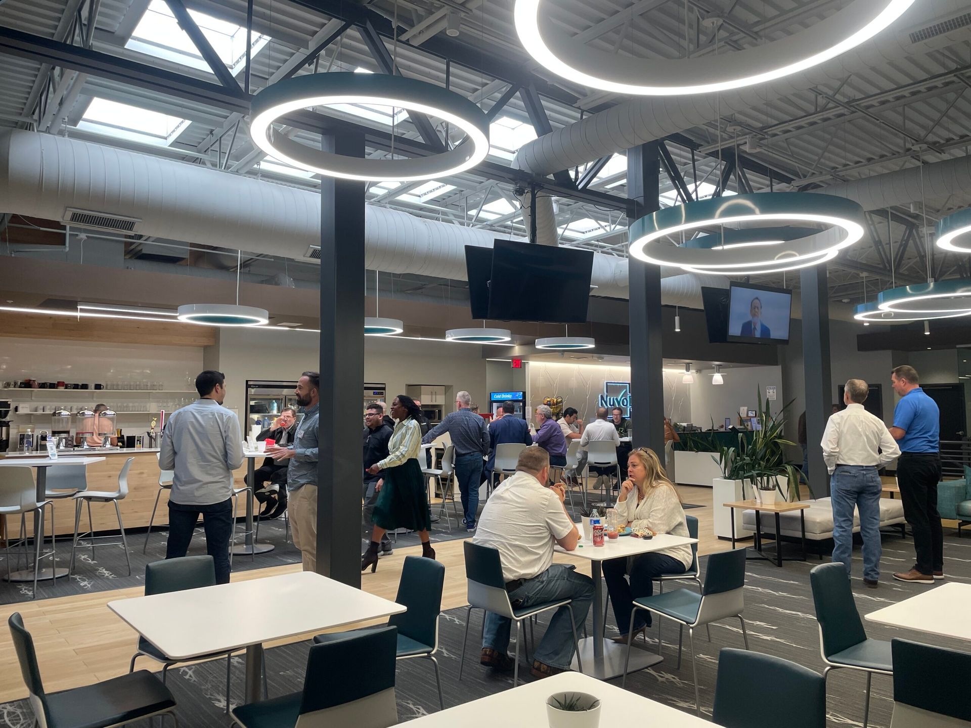 Modern cafeteria with people dining under circular ceiling lights and TV screens.