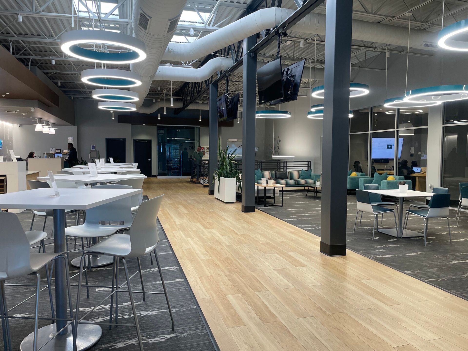 Modern open office lounge with white tables, bar seating, circular ceiling lights, and a wood walkway