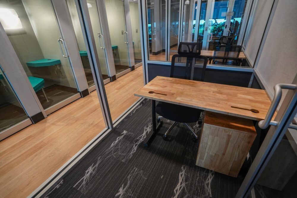 Modern office booth with a wooden desk, black chair, glass walls, and teal bench seating