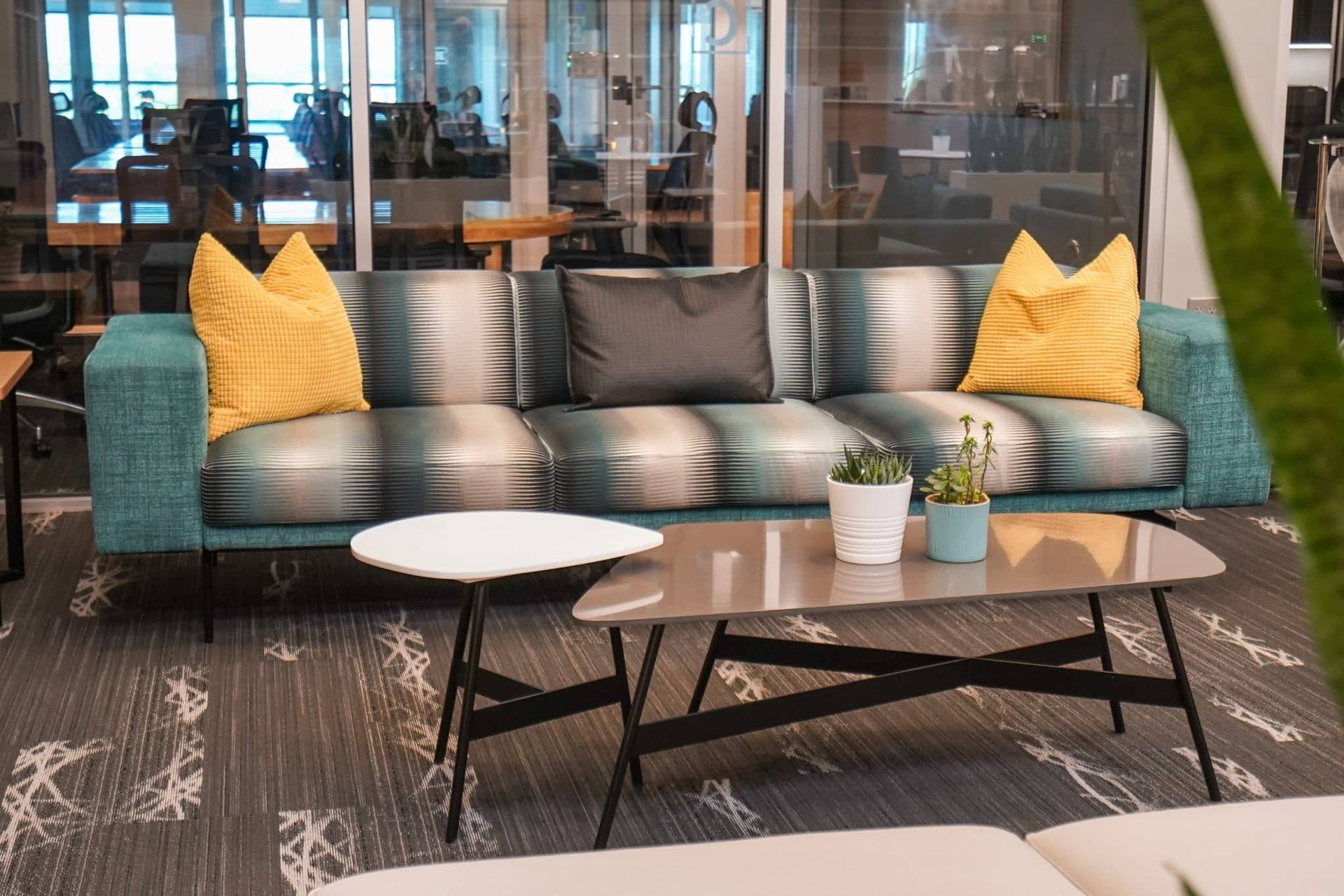 Modern lounge with teal sofa, yellow pillows, coffee tables, and office glass wall backdrop