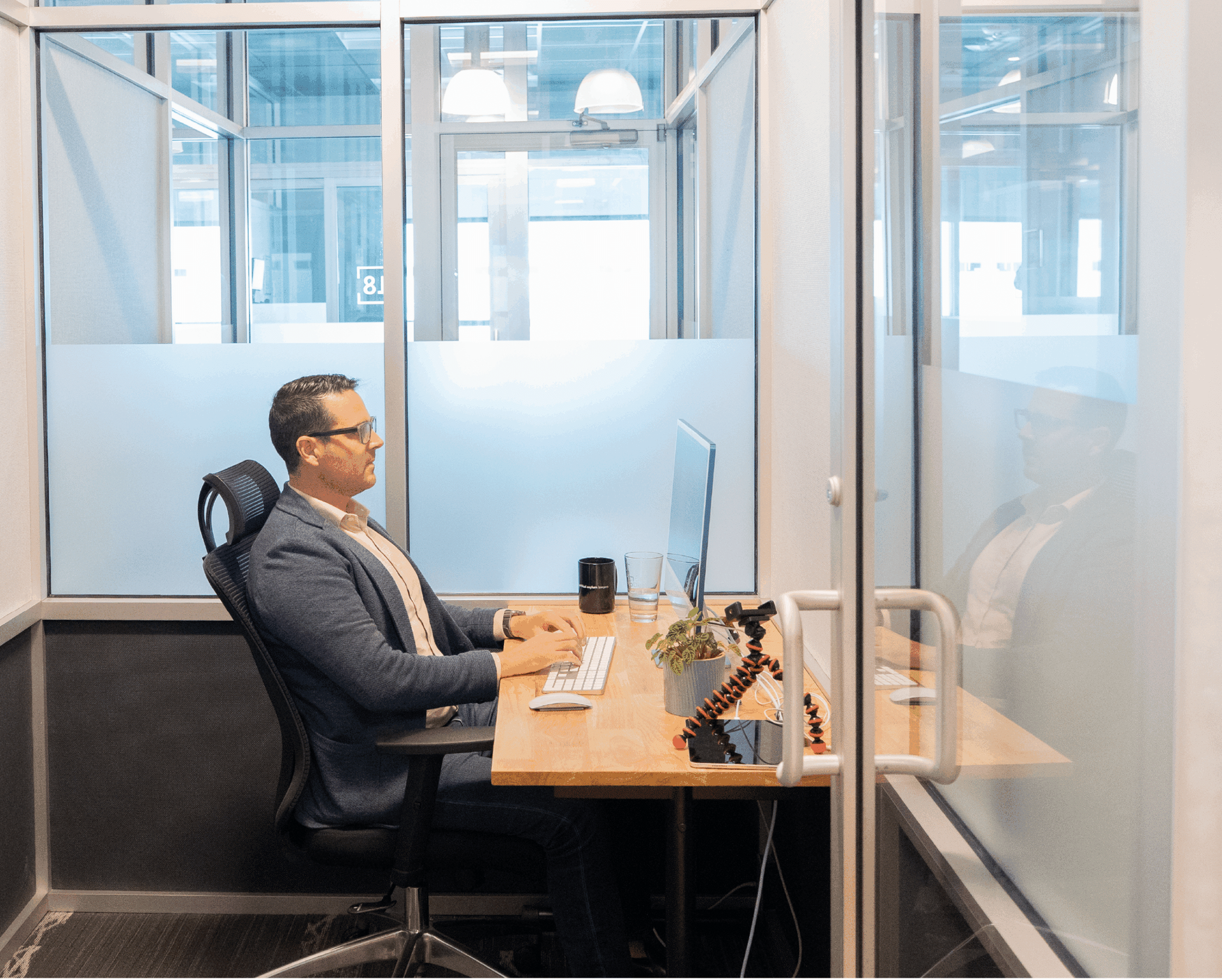 Man seated at a desk in a glass-walled office, working on a computer.