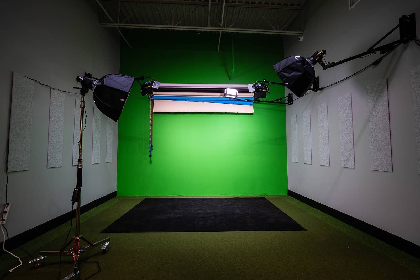 Empty indoor studio with green screen backdrop, overhead lights, and black floor mat.