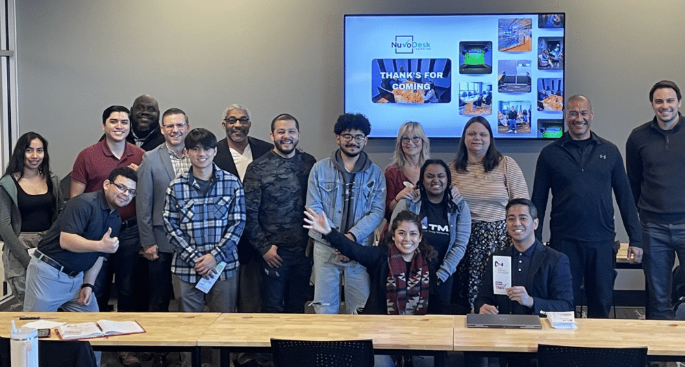 Group of people posing in a classroom, standing and sitting behind a table, with a presentation screen in the background.
