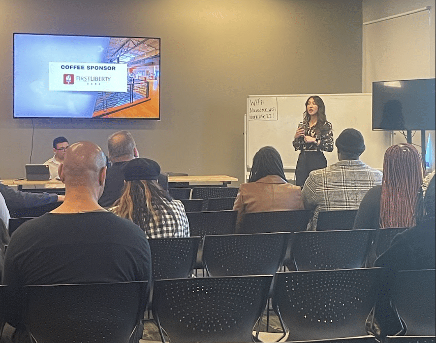 Presenter speaking to an audience in a conference room, with a slide on a wall-mounted screen.
