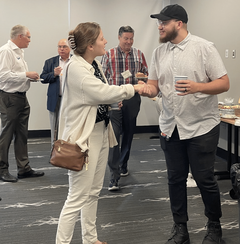 People shaking hands at an indoor networking event, with others chatting nearby.