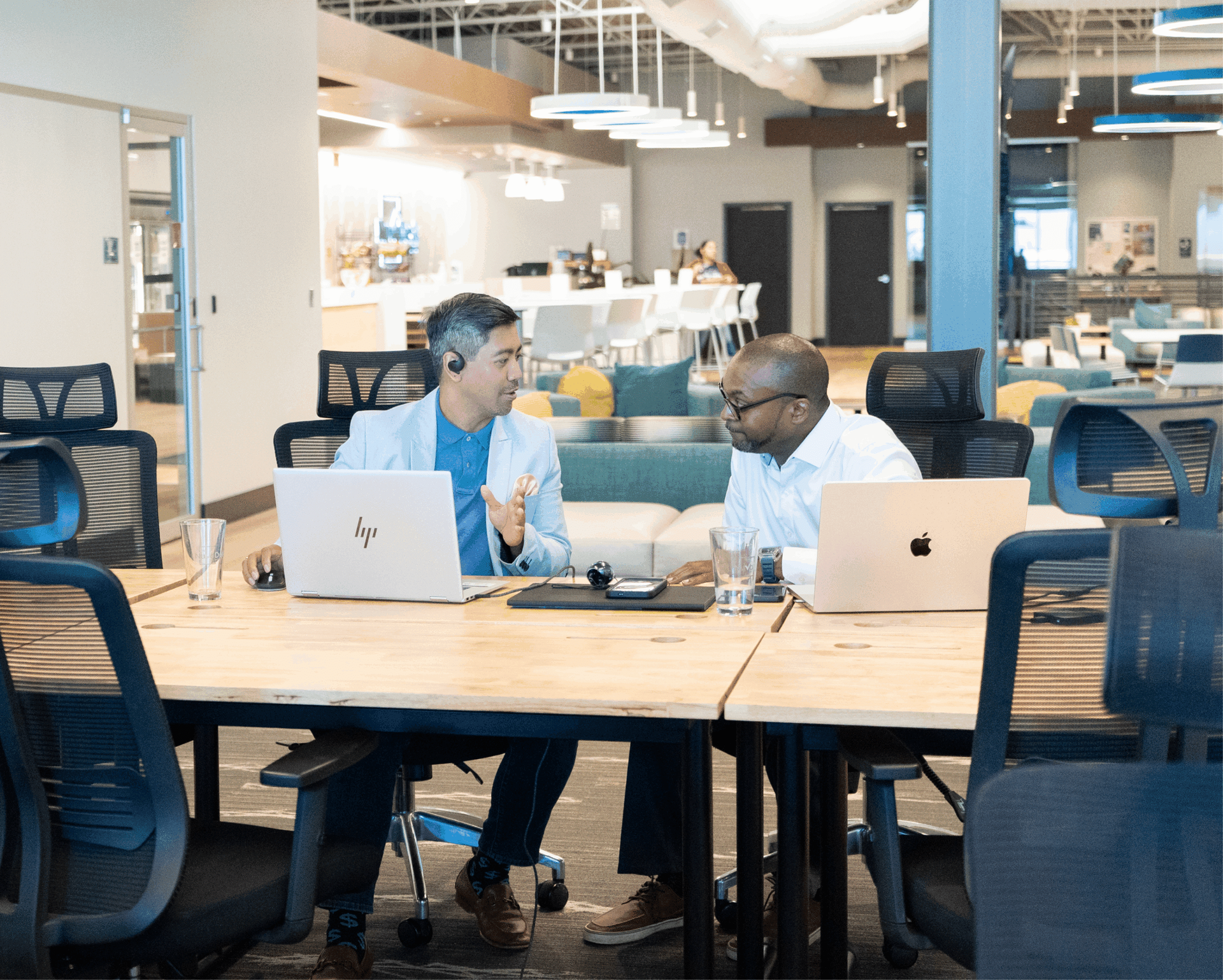 Two coworkers talking at a table with laptops in a modern office lounge.