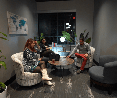 Three people seated in a modern lounge around a round table, chatting casually at night.