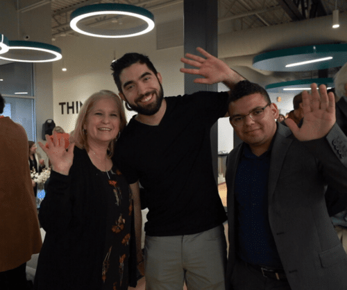 Three people smiling and waving in a modern indoor office space with circular ceiling lights