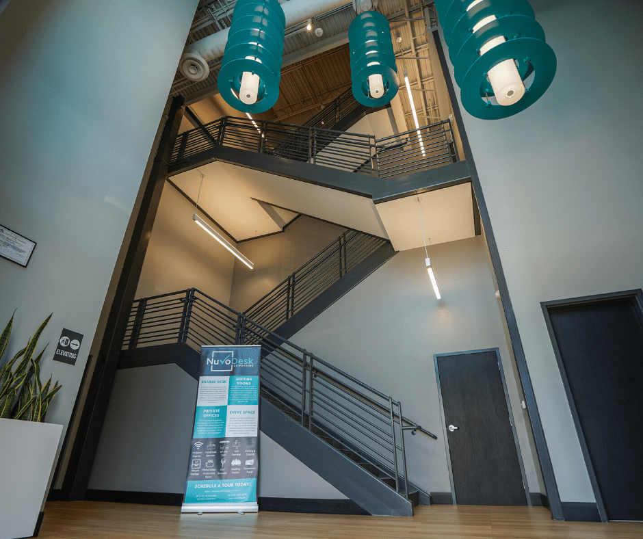 Interior stairwell with dark metal railings, teal pendant lights, and a poster stand by a wooden floor