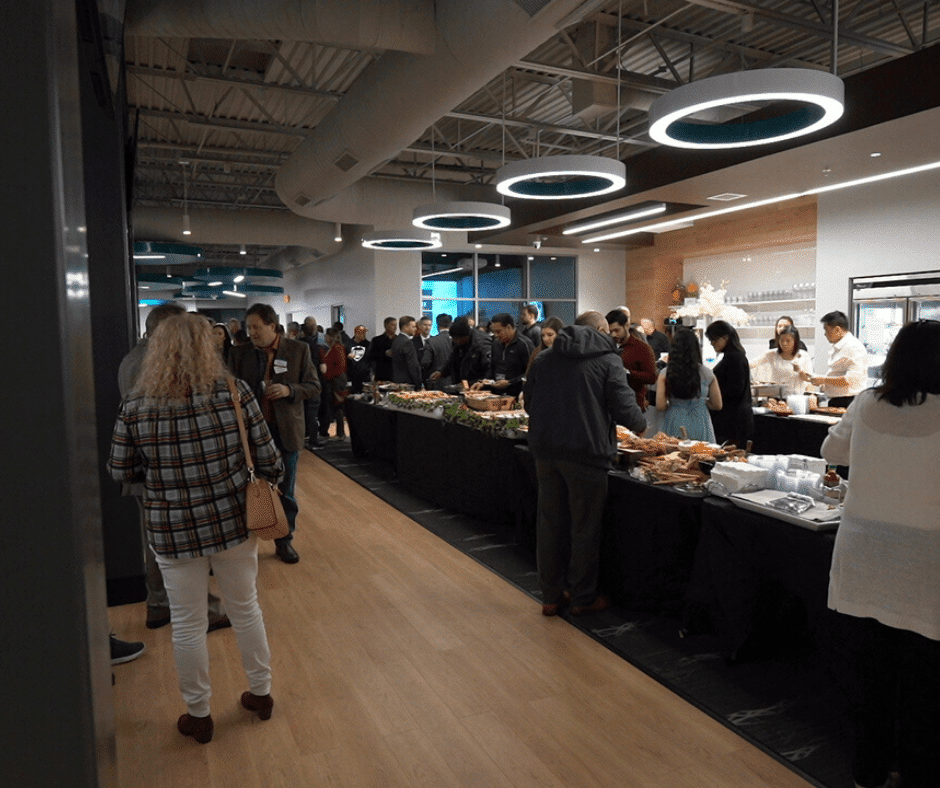 Crowded office cafeteria with people serving food at long buffet tables under modern ring lights