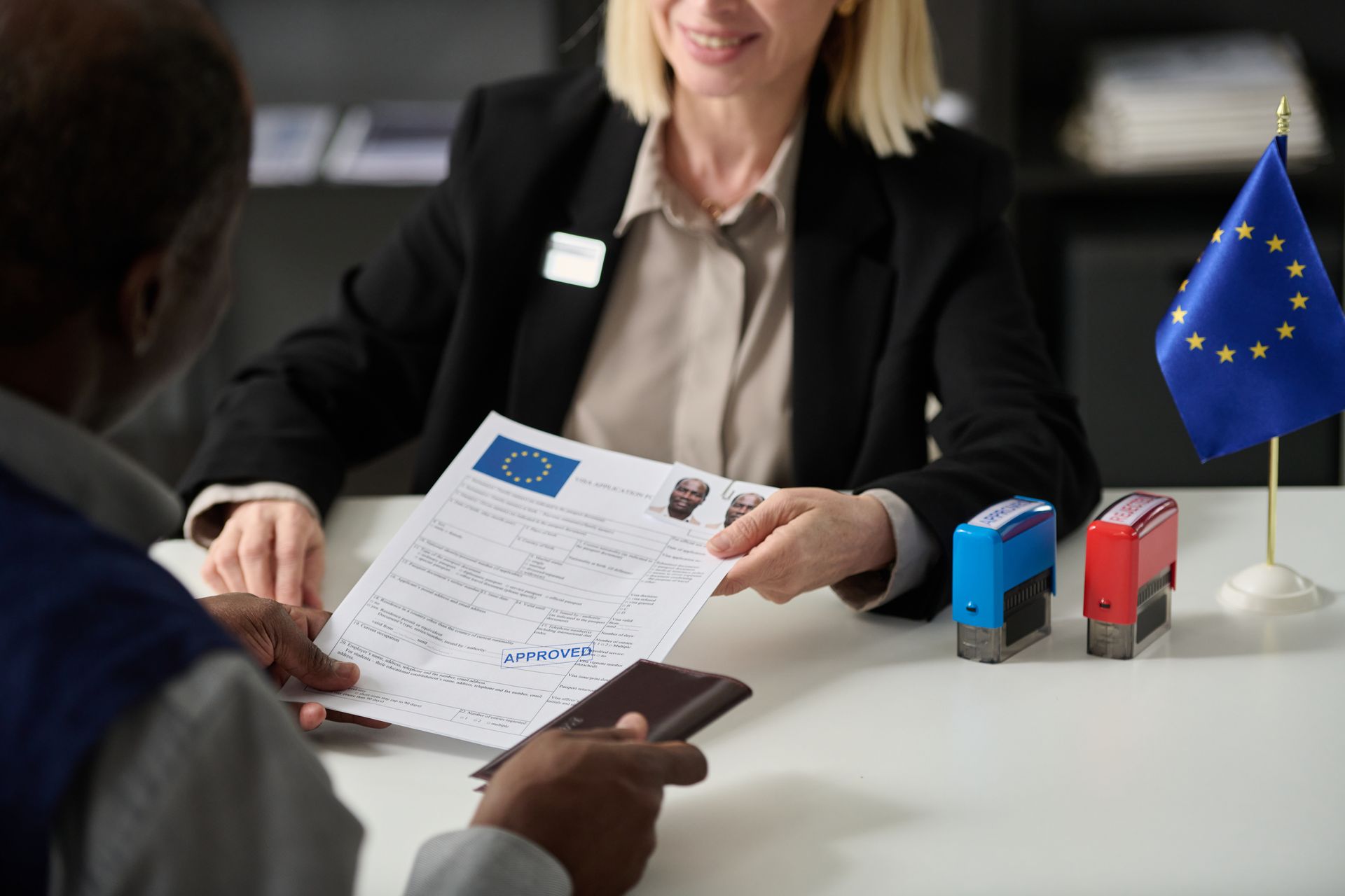 Job interview at a desk, with an applicant handing documents to an interviewer beside small desk flags.