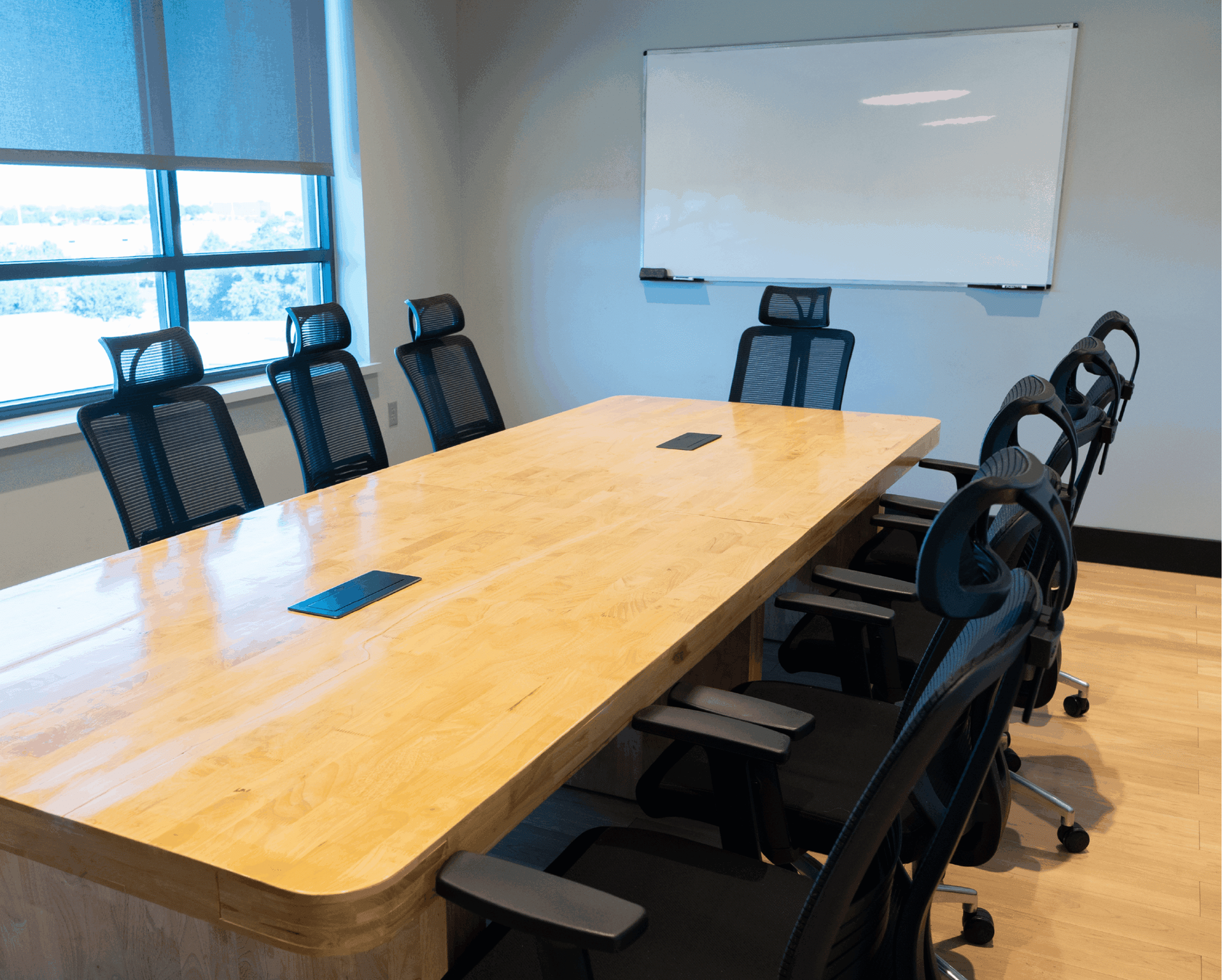 Modern conference room with a long wooden table, black chairs, and a whiteboard near windows.