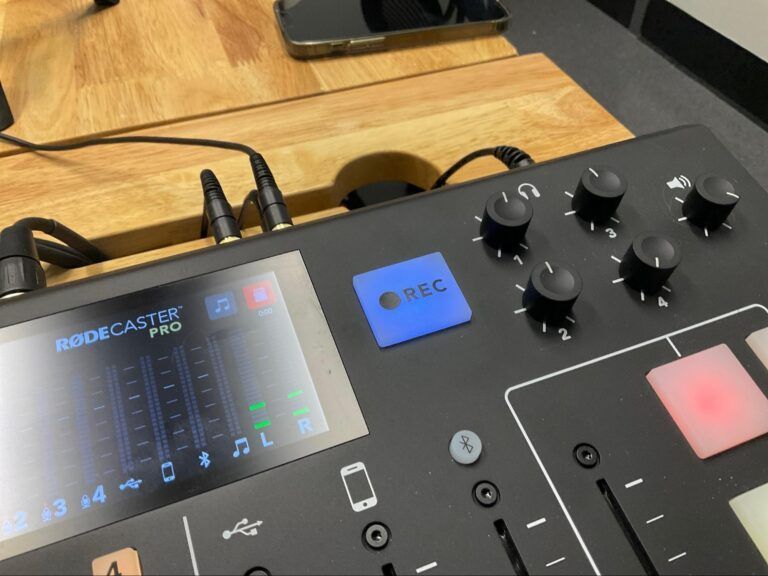 Close-up of a Rodecaster Pro audio mixer with knobs, lit display, and connected cables on a wooden desk