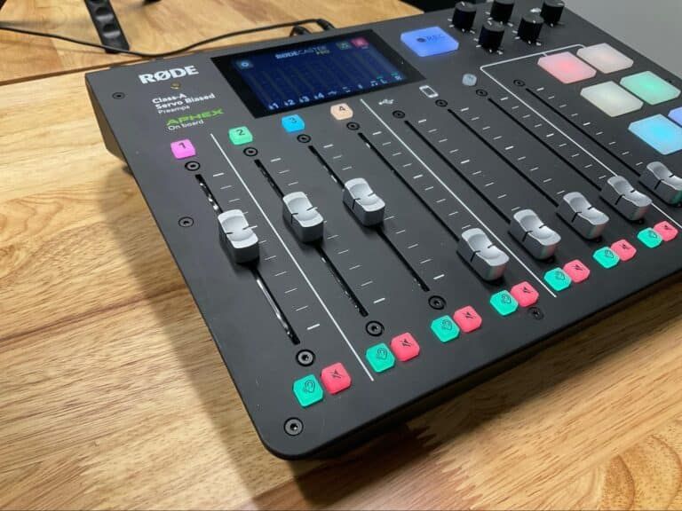 Black audio mixing console with faders, colorful buttons, and a small display on a wooden desk