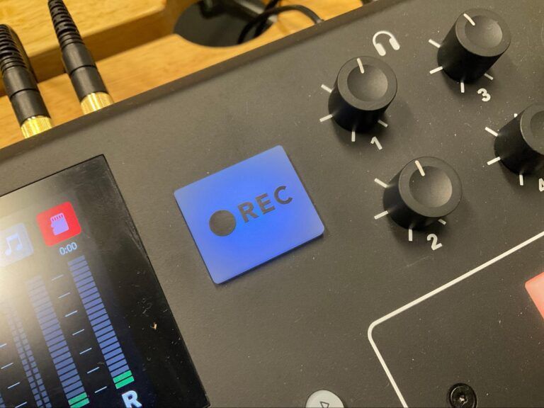 Close-up of an audio mixer with blue REC button, black knobs, and level meters lit on the left.