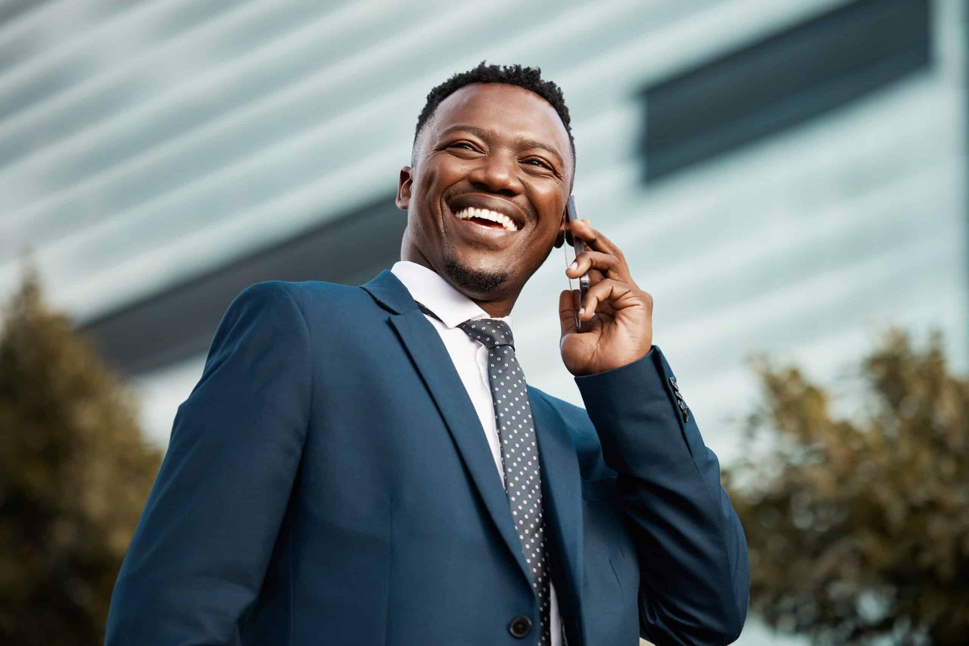 Smiling man in a blue suit talking on a cellphone outdoors near a modern building