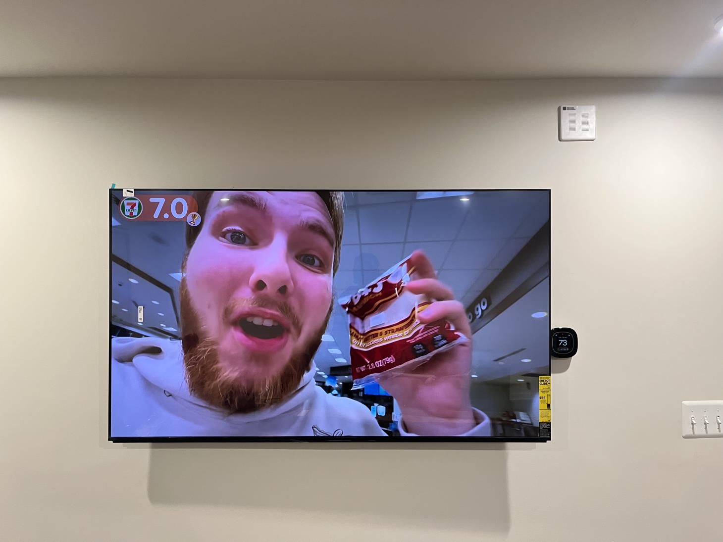 TV mounted on a wall showing a bearded man holding a dessert in a selfie-style video