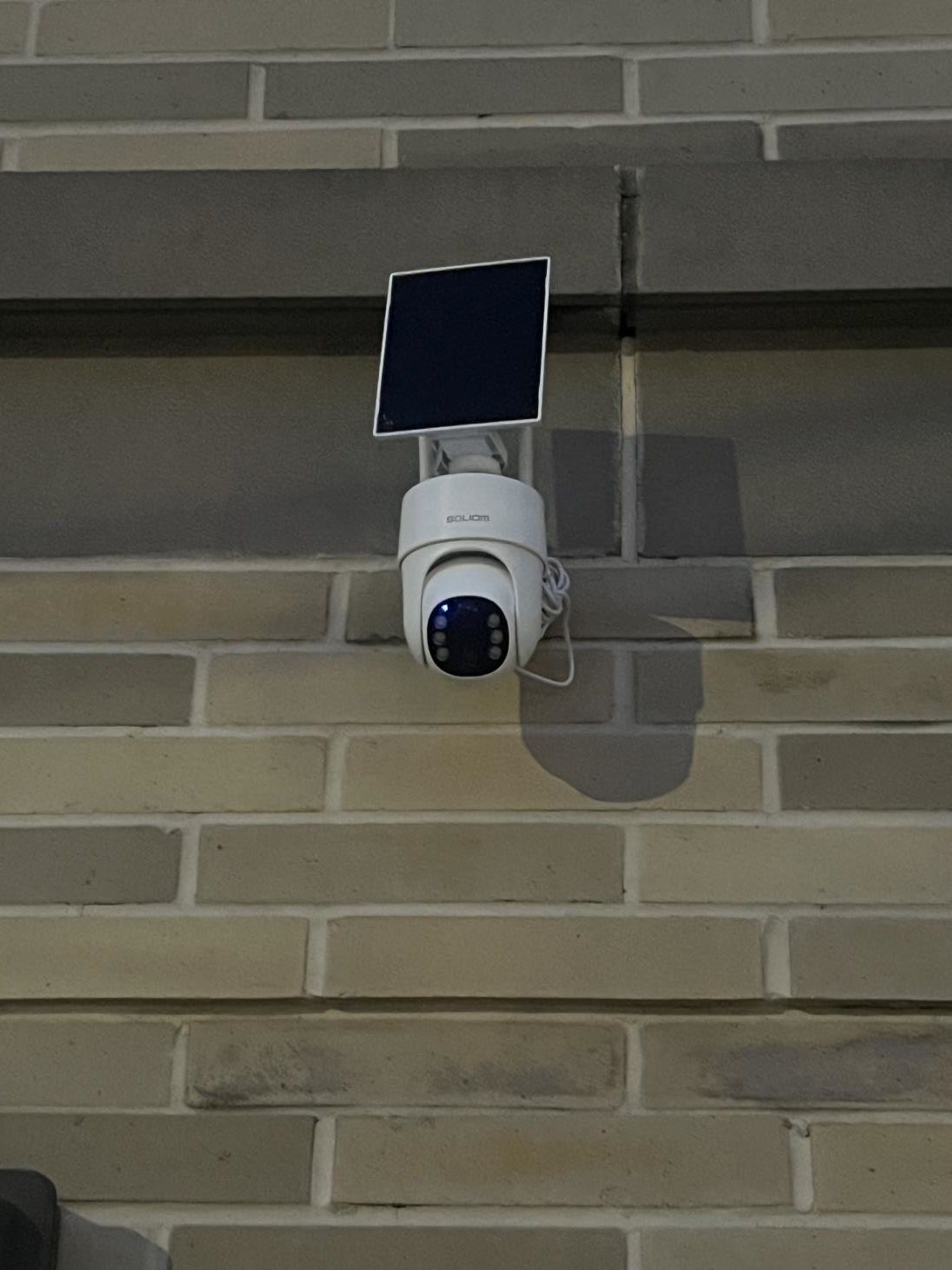 Wall-mounted white security camera with a solar panel on a brick exterior wall.