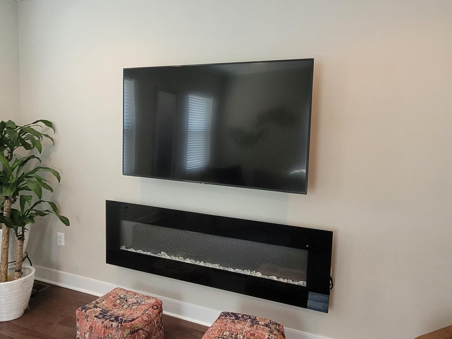 Wall-mounted TV above a black linear fireplace in a bright living room with a plant and patterned ottoman.