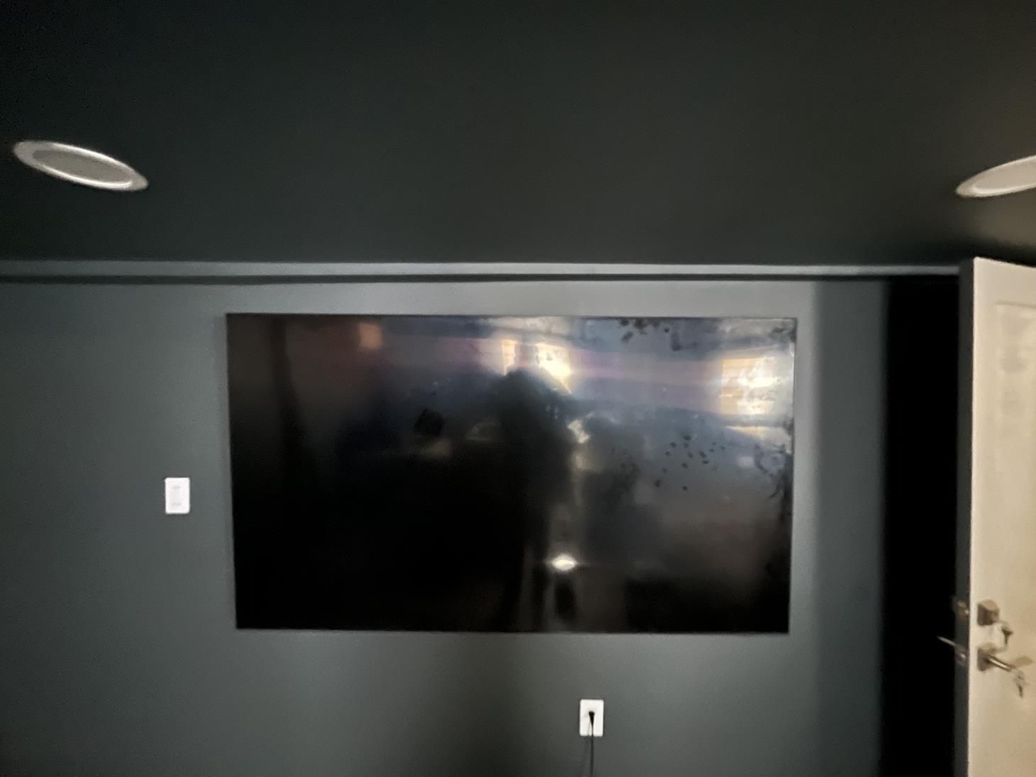 Dim room with a wall-mounted TV, dark walls, recessed lights, and an open door.