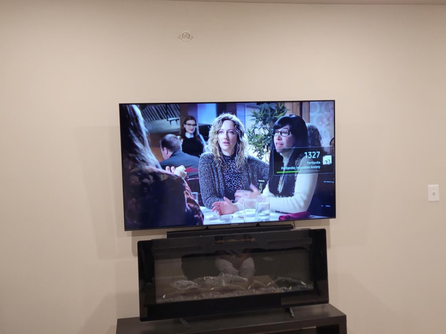TV showing a sitcom scene above a black fireplace in a beige living room