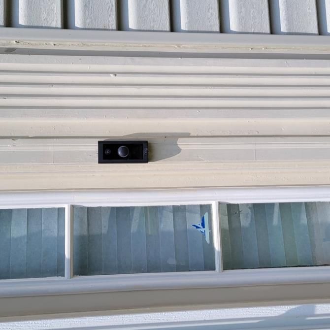 Black doorbell camera mounted above a window on a white-sided house