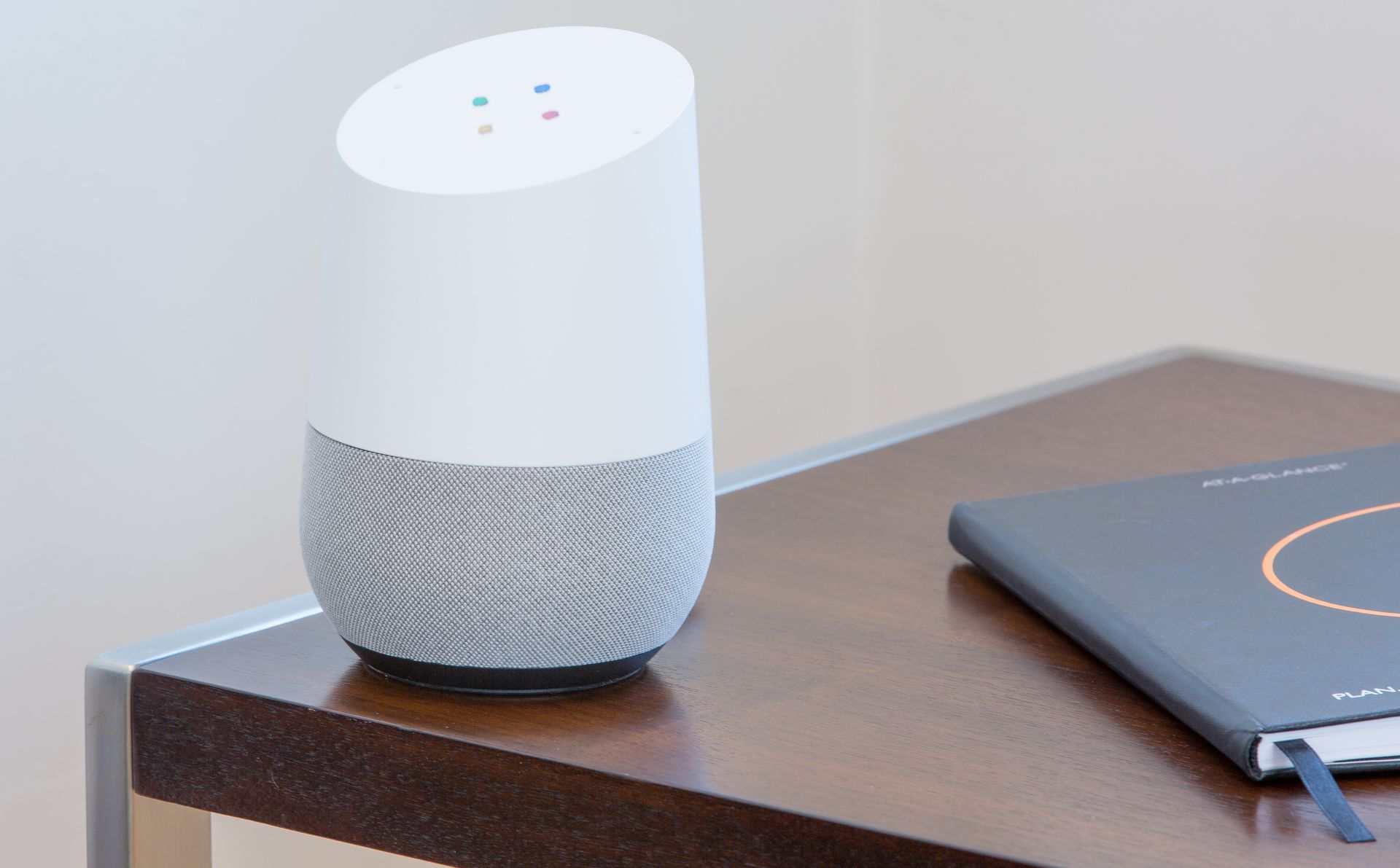 Google Home speaker on a brown nightstand next to a closed notebook; beige wall in background.