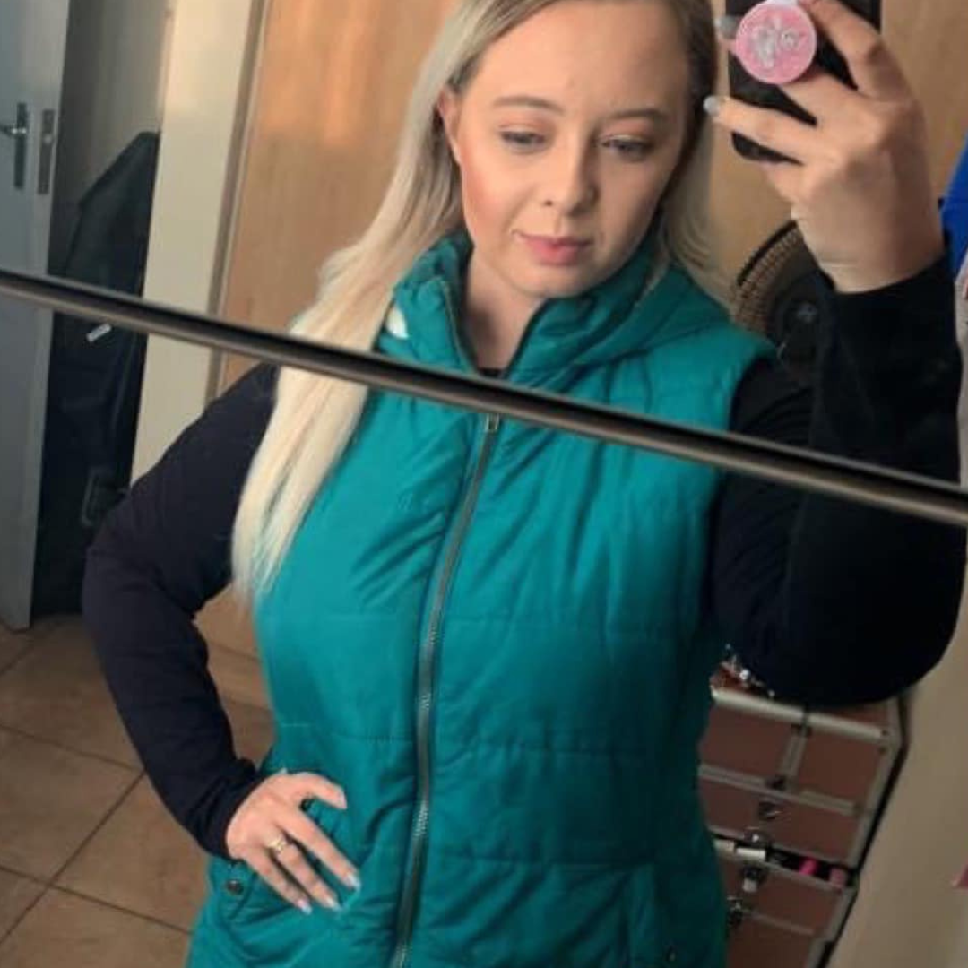 A woman in a teal vest is taking a selfie in the mirror